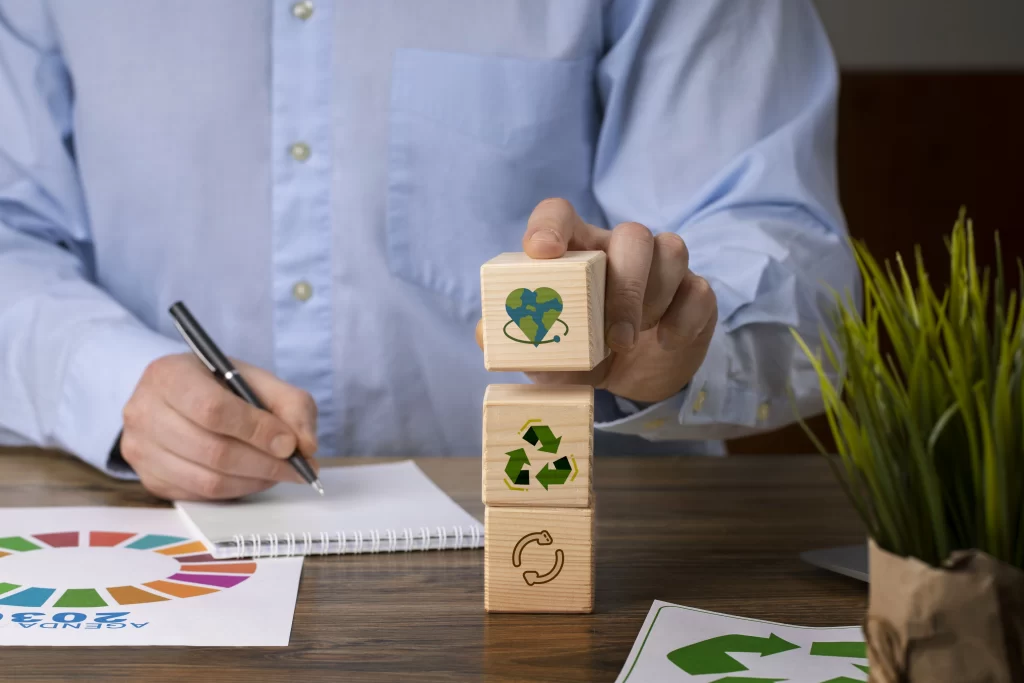 Mão de um homem empilhando blocos de madeira com ícones de sustentabilidade, representando práticas de reciclagem, proteção ao meio ambiente e economia circular, sobre uma mesa com um bloco de anotações e uma folha com o logotipo da Agenda 2030.