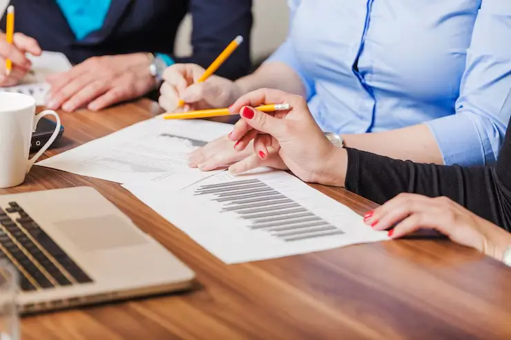 Três síndicos reunidos em uma mesa de trabalho analisam gráficos e documentos. Eles discutem questões de gestão condominial, segurando lápis e revisando dados importantes para a tomada de decisões no condomínio. No fundo, é possível ver um laptop que complementa a análise dos relatórios impressos.