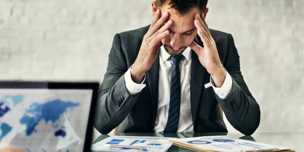 Homem de negócios sentado à mesa com as mãos na cabeça, demonstrando preocupação enquanto analisa relatórios financeiros.