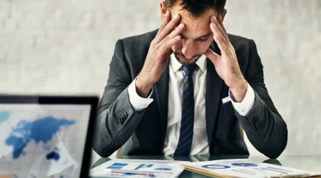 Homem de negócios sentado à mesa com as mãos na cabeça, demonstrando preocupação enquanto analisa relatórios financeiros.