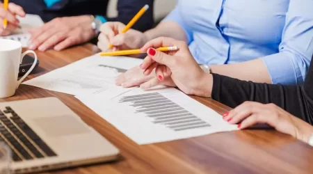 Três síndicos reunidos em uma mesa de trabalho analisam gráficos e documentos. Eles discutem questões de gestão condominial, segurando lápis e revisando dados importantes para a tomada de decisões no condomínio. No fundo, é possível ver um laptop que complementa a análise dos relatórios impressos.