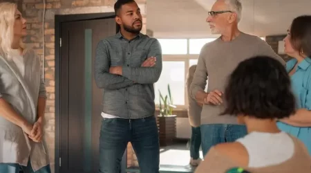 Um grupo de condôminos participando de uma reunião em um ambiente aconchegante. O grupo inclui cinco pessoas: uma mulher loira de pé à esquerda, um homem jovem com barba e braços cruzados no centro, um homem mais velho de óculos falando enquanto gesticula com as mãos, é uma mulher sentada em uma cadeira de rodas de costas para a câmera. O ambiente tem paredes de tijolos e grandes janelas que deixam entrar a luz.